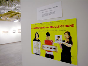 Photo of gallery space with prominent yellow poster featuring three Asian women holding different art pieces hanging on wall